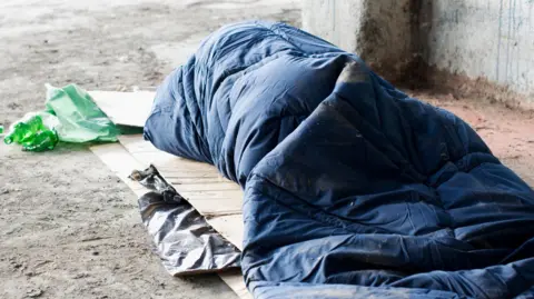A homeless person is inside a sleeping bag. They are outside and on top of cardboard. 