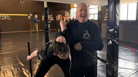Bernie holds Loughlin by the hair after besting him in a stick fight. Their shillelaghs are in both of their hands. Behind them are a couple of other participants using blue and red foam sticks.