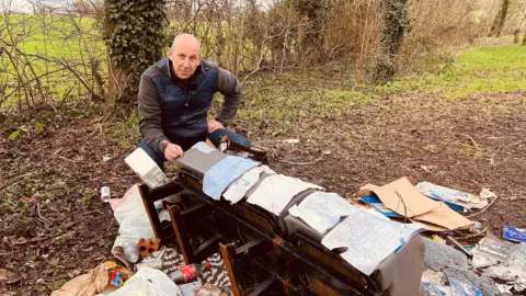 A man squatting on the edge of a field looking at a pile of rubbish that includes pieces of paper. There is a hedgerow behind him.
