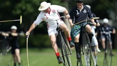 Penny Farthing Club Two players ride tall penny farthing bicycles side by side, reaching down with wooden mallets to strike a white ball on grass
