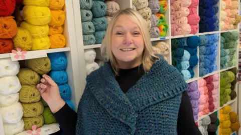 Dawn Stansfield has mid-length blonde hair and is wearing a black top and a woollen shawl. She is standing in her shop in front of a large white shelving unit filled with different coloured spools of wool.