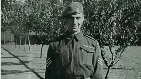 Supplied Black and white historical image of a man in Army uniform with a beret on. He is stood outdoors with a row of trees in leaf behind him.