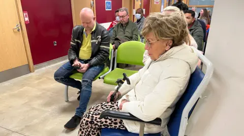 Nikki Fox/BBC At least 10 people, a mix of men and women, sitting in rows of seats in the waiting area of an urgent treatment unit. 