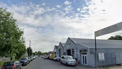 An industrial estate, featuring warehouses and parked cars. 