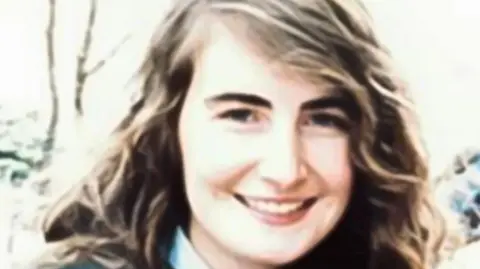 Annie McCarrick has long wavy brown hair with hints of blonde.She is wearing what appears to be a blue shirt and navy jacket. 
She is smiling and can be seen standing outside. 
A number of trees and bushes can be seen behind her. 
