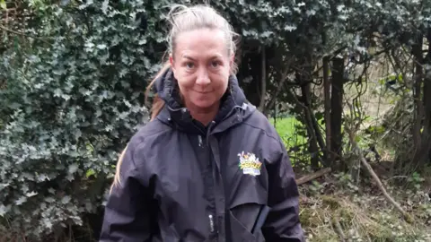 LDRS A woman wearing a black coat with her hair tied in a ponytail stands in front of a hedge