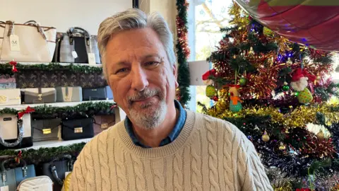 John Devine/BBC Nigel Lambert who has a short grey hair and a goatee-style grey beard. He is wearing a cream cable-knit jumper over a blue shirt. Behind him on the left are shelves with purses and bags and on his right is a Christmas tree covered in tinsel. 