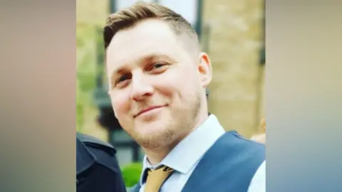 A man with short hair and a beard. He is wearing a yellow tie, a blue shirt and a navy waistcoat.