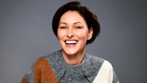Emma Willis smiling, wearing a grey, brown and white jumper