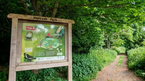 A park sign and path to the right. The area is lush and green with thick forsest.