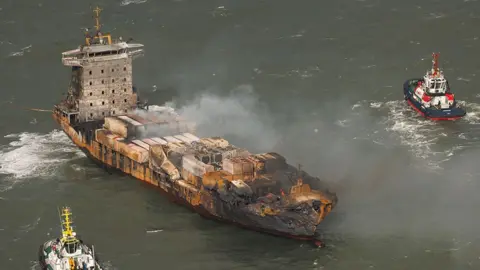 The Solong with smoke rising from its deck and fire damage to its bow and hull with two tug boats nearby