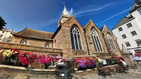 Google Maps Church with three large stained glass windows, with pink and purple flowers on the black metal fence that runs around the perimeter of the building.