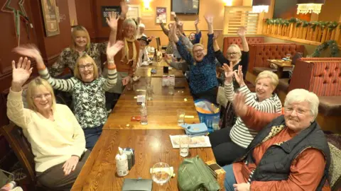 A group of women are sitting around tables in a pub. They have their hands up in the air waving to the camera. There are handbags, phones and glasses of drink on the table and they are all smiling.
