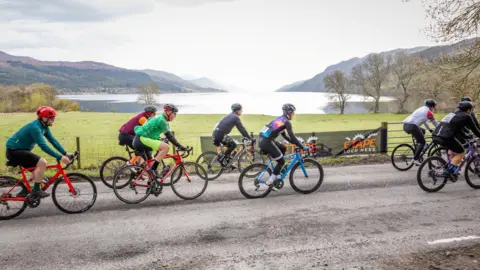 Paul Campbell Cyclist wearing different colours of cycling kit cycle along a rural road. Loch Ness is in the background.