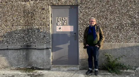 A man stands in front of a grey and brown pebbedashed wall, next to a grey metal door with a warning notice on it. He has grey hair and is wearing glasses, a dark green jacket over a dark top and blue scarf, blue jeans with a turn-up and black shoes.