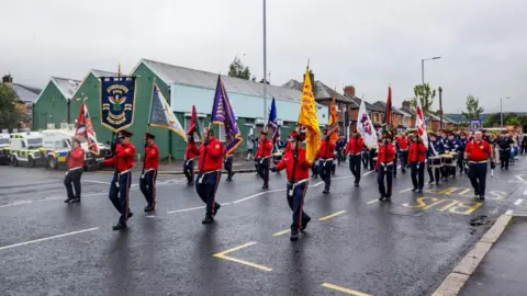 A flute band wearing red shirts and navy trousers. The front group are holding flags. Drummers can be seen in the background. Police land rovers are parked alongside the band.