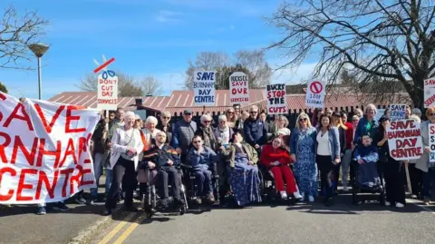 Hazel Howe A group of people, some in wheelchairs, sitting in front of a single-storey day centre building. Some are holding "Save Burnham Day Centre" placards. There is a large white banner to the left with "Save Burnham Centre" in red capitals.