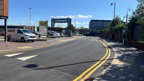 BBC A street with double yellow lines runs past a car park which has a large green and red sign. Several cars are parked on the site, the Hull tidal barrier and modern buildings are visible in the background.