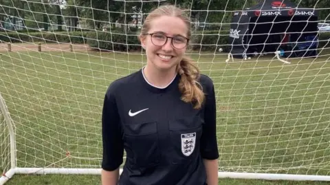 BBC A teenage girl with blonde hair in two plaits, wearing glasses and a black referee uniform, stood in front of a goal on a football pitch