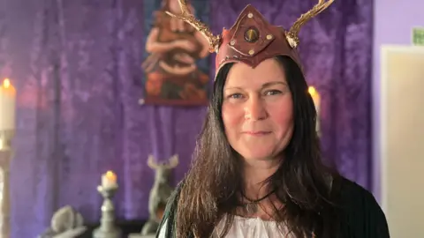Angela smiles at the camera. She wears a headdress made from brown leather with a stone in the middle and gold antlers. She stands in front of a purple altar.