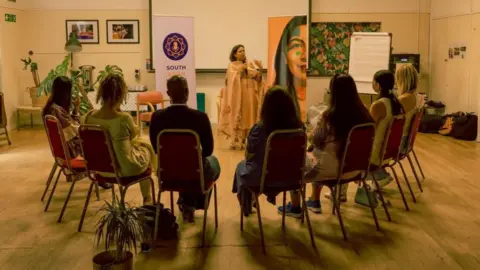 Kamil Hendrych South Asian Voices Bristol CIC hosting a group discussion in a function room in Bristol. One member is speaking using a microphone. Seven other people are listening while seated in a semi circle. The woman speaking is wearing a sari. 