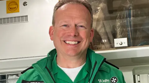 Julia Gregory/BBC A smiley man stands in an ambulance wearing a green uniform 