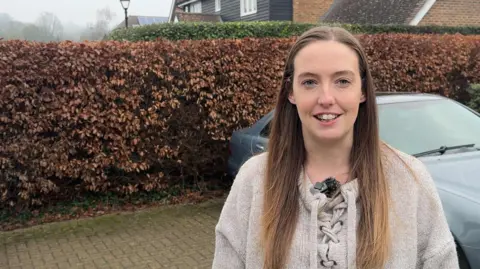 A woman with long brown hair and cream coloured jumper stands in front of a hedge. 