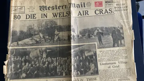 A copy of the Western Mail newspaper, dated 13th March 1950 with the headline '80 die in Welsh air crash', alongside a photo of the wreckage and another photo showing the passengers in front of the plane, taken before they left for Ireland.