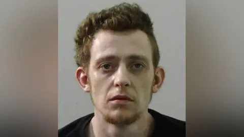 Northumbria Police Mugshot of Reece Galbraith. He is gaunt and pale with thick ginger hair and a ginger goatee, wearing a black t shirt
