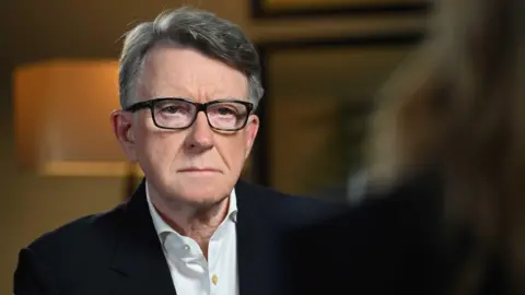 BBC Close up of Peter Mandelson wearing thick black frames, a dark jacket and white shirt.