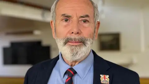 A man with a beard in a jacket and tie looks towards the camera