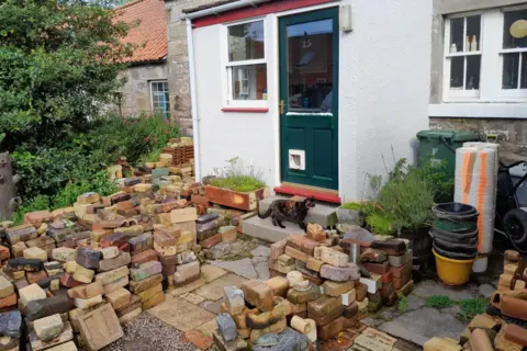 Ian Suddaby Ian Suddaby's garden is covered in piles of colourful bricks. There is a cat on his doorstep and his door is green and has a glass pane in it,