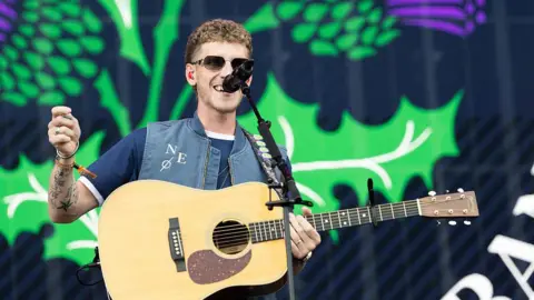 Getty Images Nathan Evans on a stage in front of a microphone and playing the guitar. He is wearing a blue t-shirt, denim gilet and dark sunglasses. A thistle is projected on a screen in the background.