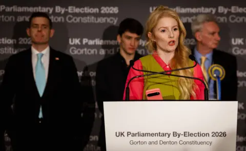 LightRocket via Getty Images Hannah Spencer looks to the right of frame as she speaks while standing at a lecturn that reads 'UK parliamentary by-election 2026; Gorton and Denton Constituency', with three other candidates standing onstage behind her.