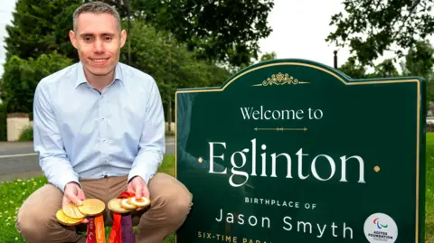 Sprinter Jason Smyth kneels beside a new welcome sign in the village where he was born. He is wearing a blue shirt and beige trousers and is holding six gold medals. He is smiling and has short greying hair.