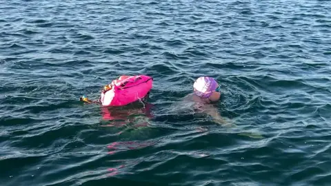 A woman wearing goggles and a purple swimming hat and pink toe float following behind swimming in the lake.