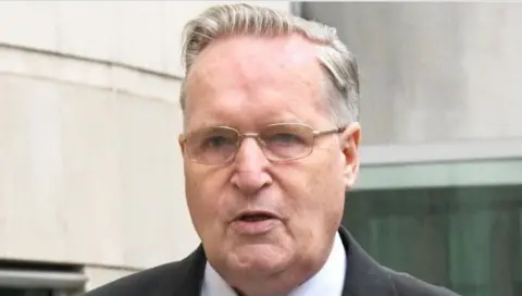 Alan Lewis/ PHOTOPRESS A white haired man wearing gold framed glasses in a grey suit, white shirt and black patterned tie