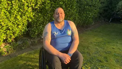 Mike Ryan, a man with tattoos on his arms, he wears a blue vest with the three legs of man and smiles as he sits in the garden in his wheelchair.