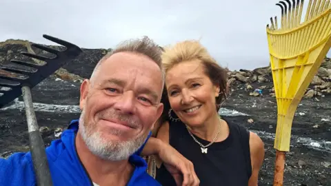 Dean Ward and Allie Evans. He has grey hair and a beard and is wearing a blue top. She has a black top, silver earrings and a silver necklace with butterfly pendant. They are holding rakes and stood on a blackened mountain