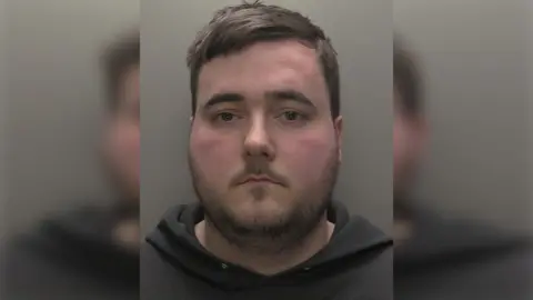Custody photograph of Connor Laud he has short brown hair and beard and is wearing a dark hooded top