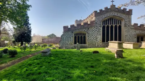 Outside Ewelme Church on a sunny day. It has a distinctive chequered pattern made with knapped flint and limestone ashlar. It is a sunny day. Many graves lay in its churchyard.