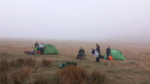 Mount Kelly College A group of people putting up green tents and setting up camp in the middle of the moor.