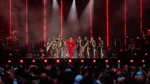 Daniel Ramos Bruno Mars performs on stage at Wembley stadium with his backing band, The Hooligans. The Hooligans wear black suits with gold details, Bruno Mars wears an all red suit.