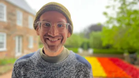 A mid shot of Stuart Temple. He is wearing a grey jumper, glasses, and a dark yellow woolly hat.