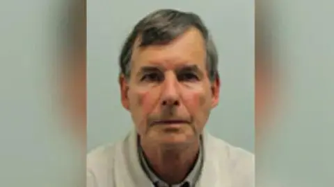 A police mugshot of a man with grey head and wearing a white jacket.