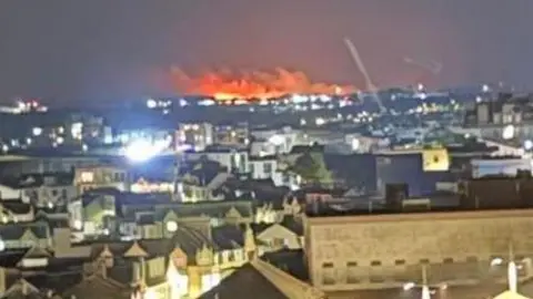 A large moorland fire burns in the distance from Newquay. Large orange flames head towards the night sky. Hundreds of buildings are in the foreground.