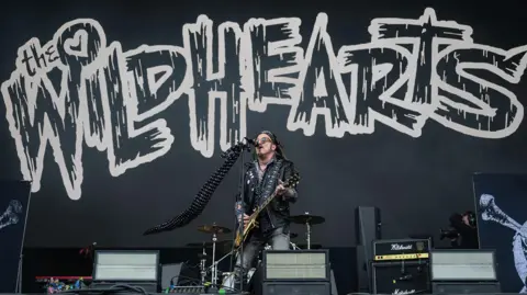 Getty Images A man in a black leather jacket, playing guitar on stage. He is singing into a microphone. Behind him is a black backdrop, with "The Wildhearts" written in white letters.