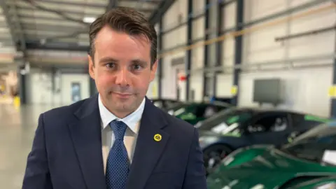 Alex Dunlop/BBC Matt Nice standing in front of a row of green Lotus produced cars. He is looking directly at the camera and smiling and is wearing a white shirt, navy suit jacket and navy tie. On the lapel of his suit is a yellow Lotus pin badge.