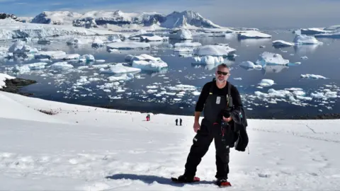 Man standing on snow in black outfit with snow-covered water in background behind him