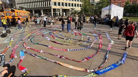 Aaron Outram/BBC A chain of many different coloured football scarfs placed in a spiral shape on the floor of a town square